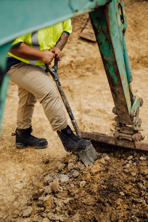 Person wearing Oryx boots, using a shovel at a construction site with excavator equipment in the background.